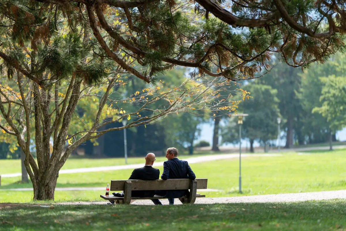 Zwei Männer im Anzug sitzen auf einer Parkbank unter einem Baum, im Hintergrund der Traunsee