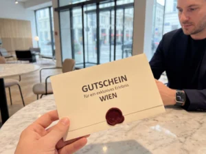 Eine Hand hält einen versiegelten Gutschein mit der Aufschrift „Gutschein für ein exklusives Erlebnis Wien“. Der Umschlag ist mit rotem Wachssiegel verschlossen, im Hintergrund sitzt eine Person an einem Tisch in einem modernen Café.