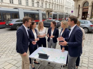 Gruppe von sechs Agenten in Businesskleidung steht um einen Tisch mit Stadtplan und Tablet in der Wiener Innenstadt, im Hintergrund Straßenbahn und historische Gebäude nahe Café Central.